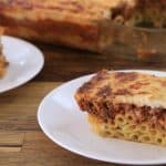 A slice of baked pasta with a meat and cheese topping is placed on a white plate. The slice appears to have layers of pasta, meat sauce, and a cheese crust. In the background, there is a glass baking dish containing the remaining portions of the baked pasta.