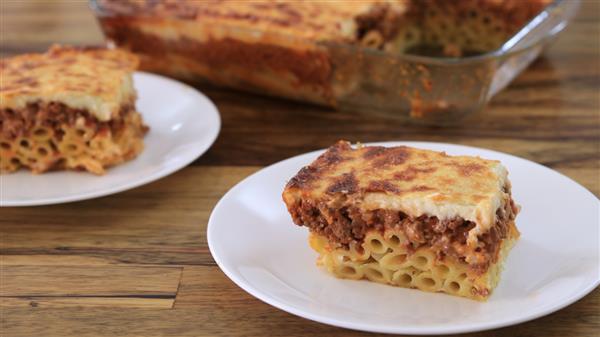 A slice of baked pasta with a meat and cheese topping is placed on a white plate. The slice appears to have layers of pasta, meat sauce, and a cheese crust. In the background, there is a glass baking dish containing the remaining portions of the baked pasta.