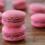 A pair of pink macarons is stacked on a wooden surface, with more macarons blurred in the background. Each macaron has a smooth, glossy filling sandwiched between two delicate, light pink shells.