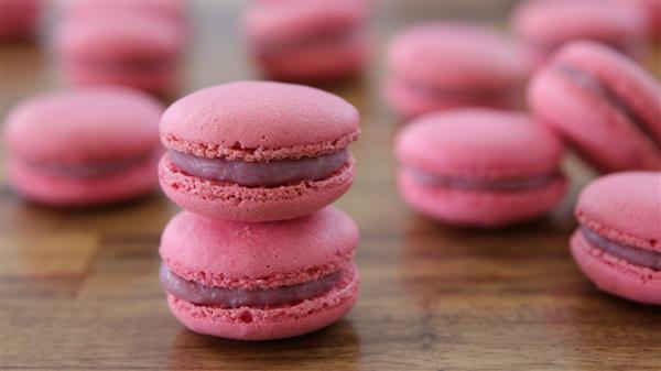 A pair of pink macarons is stacked on a wooden surface, with more macarons blurred in the background. Each macaron has a smooth, glossy filling sandwiched between two delicate, light pink shells.