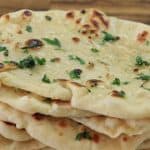 A stack of freshly baked naan bread topped with melted butter, finely chopped garlic, and sprinkled with fresh parsley. The naans are slightly golden with some charred spots, resting on a wooden surface.