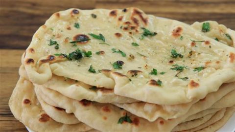 A stack of freshly baked naan bread topped with melted butter, finely chopped garlic, and sprinkled with fresh parsley. The naans are slightly golden with some charred spots, resting on a wooden surface.