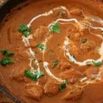 A close-up of a dish of butter chicken garnished with fresh cilantro and a swirl of cream, served in a black bowl. A side of naan bread, adorned with herbs and garlic, is partially visible to the left of the bowl on a wooden surface.