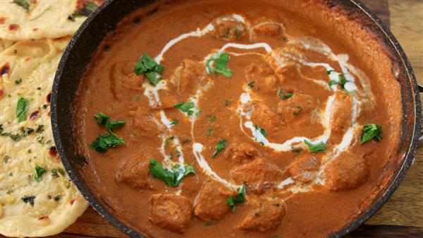 A close-up of a dish of butter chicken garnished with fresh cilantro and a swirl of cream, served in a black bowl. A side of naan bread, adorned with herbs and garlic, is partially visible to the left of the bowl on a wooden surface.