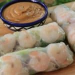 Fresh spring rolls with visible shrimp are placed on a wooden board, along with mint leaves. A small bowl of peanut dipping sauce is positioned behind the rolls. The translucent rice paper wrappers showcase the colorful vegetables and noodles inside.