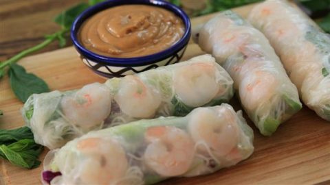 Fresh spring rolls with visible shrimp are placed on a wooden board, along with mint leaves. A small bowl of peanut dipping sauce is positioned behind the rolls. The translucent rice paper wrappers showcase the colorful vegetables and noodles inside.