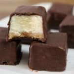 A close-up view of chocolate-coated cheesecake bites on a white plate. One of the bites has a piece removed, revealing the creamy cheesecake filling and a thin layer of crust. Other bites are visible in the background, all coated in smooth chocolate.