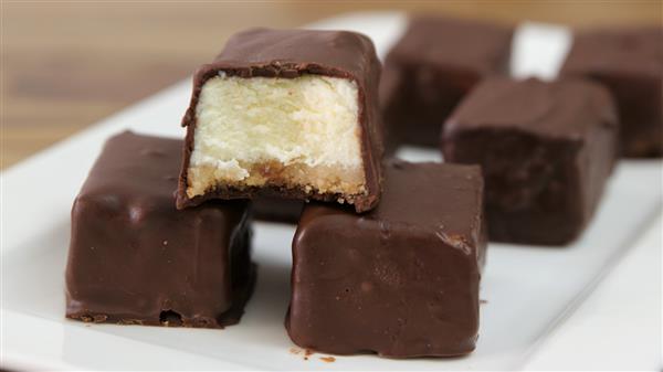 A close-up view of chocolate-coated cheesecake bites on a white plate. One of the bites has a piece removed, revealing the creamy cheesecake filling and a thin layer of crust. Other bites are visible in the background, all coated in smooth chocolate.