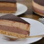 A slice of layered chocolate and caramel mousse cake on a white plate, with a crumbly base and a glossy chocolate topping. Two forks are placed on the plate, and another slice of the cake is visible in the background.