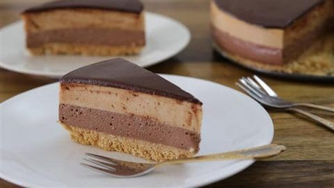 A slice of layered chocolate and caramel mousse cake on a white plate, with a crumbly base and a glossy chocolate topping. Two forks are placed on the plate, and another slice of the cake is visible in the background.