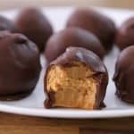 A white plate holds several chocolate-covered peanut butter balls. One of the balls in the foreground has a bite taken out, revealing the creamy peanut butter filling inside. The plate sits on a wooden surface.