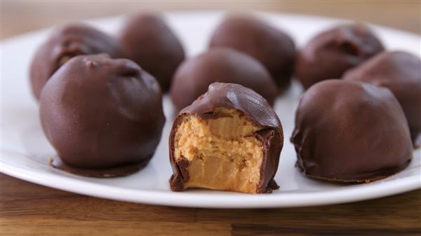 A white plate holds several chocolate-covered peanut butter balls. One of the balls in the foreground has a bite taken out, revealing the creamy peanut butter filling inside. The plate sits on a wooden surface.