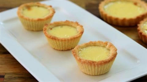 Four golden-brown egg tarts are neatly arranged on a rectangular white plate. The tarts have a crisp, flaky crust and a smooth, custard-like filling. The plate is placed on a wooden surface, giving a warm and inviting appearance.