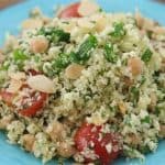 A vibrant plate of cauliflower rice salad is served. The dish includes finely chopped cauliflower, halved cherry tomatoes, chickpeas, and green herbs, all topped with sliced almonds. The ingredients are fresh, colorful, and arranged on a blue plate.