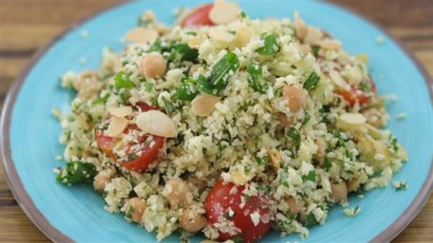 A vibrant plate of cauliflower rice salad is served. The dish includes finely chopped cauliflower, halved cherry tomatoes, chickpeas, and green herbs, all topped with sliced almonds. The ingredients are fresh, colorful, and arranged on a blue plate.