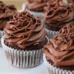 A plate of chocolate cupcakes, each topped with a generous swirl of creamy chocolate frosting, displayed in white paper liners. The cupcakes are arranged closely together in an appealing manner. The background is softly blurred, emphasizing the cupcakes.