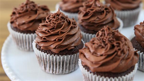 A plate of chocolate cupcakes, each topped with a generous swirl of creamy chocolate frosting, displayed in white paper liners. The cupcakes are arranged closely together in an appealing manner. The background is softly blurred, emphasizing the cupcakes.