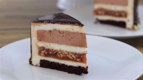 A slice of layered cake on a white plate, featuring a bottom layer of dark chocolate cake, a layer with nuts and caramel, two light cream layers with a chocolate mousse-like layer in between, and a glossy chocolate glaze on top. Another slice is visible in the background.