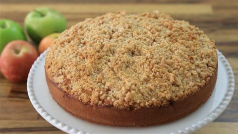 A round apple crumb cake sits on a white plate, featuring a crumbly, golden-brown topping. In the background, there are green and red apples placed on a wooden surface.