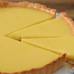 A close-up of a lemon tart with a smooth, bright yellow filling. The tart has a golden-brown outer crust and is set on a white plate. Three triangular slices are cut and arranged neatly on the plate. The background is a blurred wood surface.