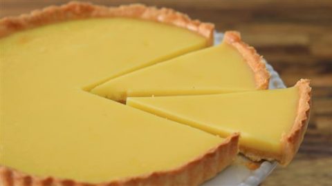 A close-up of a lemon tart with a smooth, bright yellow filling. The tart has a golden-brown outer crust and is set on a white plate. Three triangular slices are cut and arranged neatly on the plate. The background is a blurred wood surface.