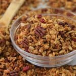 A clear glass bowl filled with granola sits on a surface scattered with more granola. The granola is a mix of toasted oats, pecans, and dried cranberries. A wooden spoon is also visible, partially covered with granola.