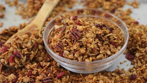 A clear glass bowl filled with granola sits on a surface scattered with more granola. The granola is a mix of toasted oats, pecans, and dried cranberries. A wooden spoon is also visible, partially covered with granola.