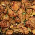 A baking dish filled with roasted chicken drumsticks, thighs, and quartered potatoes, garnished with sprigs of rosemary. The chicken and potatoes are golden brown and lightly seasoned, with caramelized onions scattered throughout.