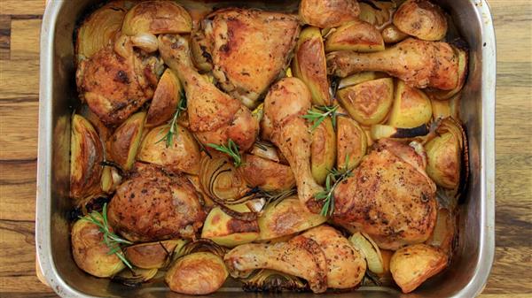 A baking dish filled with roasted chicken drumsticks, thighs, and quartered potatoes, garnished with sprigs of rosemary. The chicken and potatoes are golden brown and lightly seasoned, with caramelized onions scattered throughout.