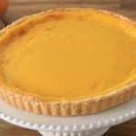 A freshly baked, golden yellow, lemon tart sits on a white cake stand with a fluted edge. The tart has a smooth, glossy surface with a golden brown crust. In the background, there are a couple of apples and an orange resting on a wooden table.