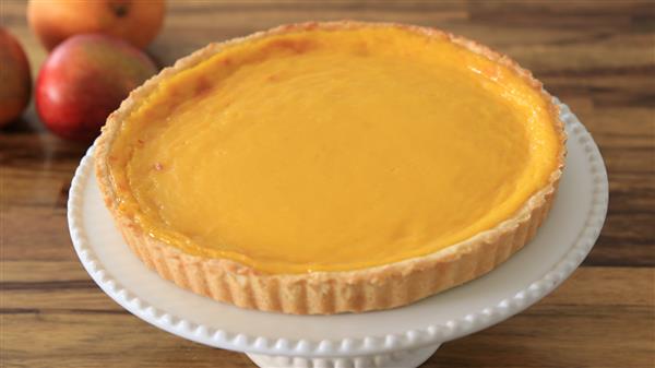 A freshly baked, golden yellow, lemon tart sits on a white cake stand with a fluted edge. The tart has a smooth, glossy surface with a golden brown crust. In the background, there are a couple of apples and an orange resting on a wooden table.