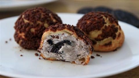 Three cream puffs sit on a white plate. Two are whole, while one is cut in half, revealing cookies and cream filling inside. The puffs have a crispy choux pastry exterior lightly dusted with chocolate crumbs, and bits of cookie are visible in the filling.