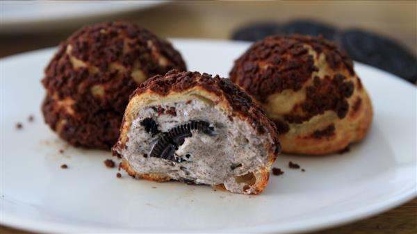 Three cream puffs sit on a white plate. Two are whole, while one is cut in half, revealing cookies and cream filling inside. The puffs have a crispy choux pastry exterior lightly dusted with chocolate crumbs, and bits of cookie are visible in the filling.