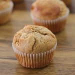 Close-up image of several muffins arranged on a wooden surface. The muffins have a golden-brown top with a slightly cracked surface and are held in white paper liners. The background is out of focus but shows more muffins.