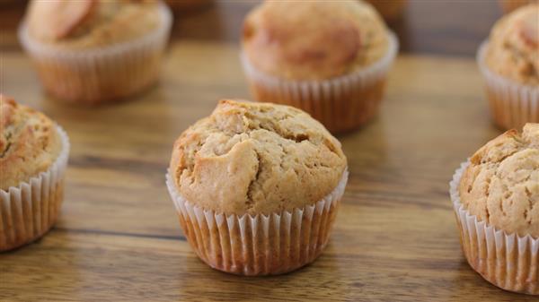 Close-up image of several muffins arranged on a wooden surface. The muffins have a golden-brown top with a slightly cracked surface and are held in white paper liners. The background is out of focus but shows more muffins.