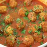 A white dish filled with meatballs covered in a rich, red tomato sauce. The meatballs are garnished with chopped green herbs, likely parsley or cilantro, and the dish sits on a wooden surface.