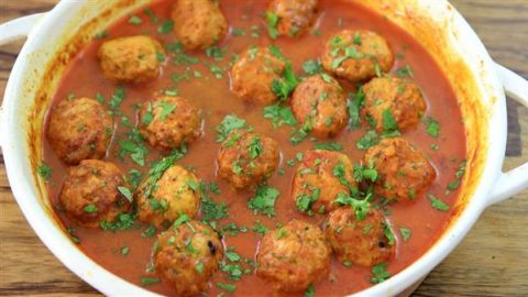 A white dish filled with meatballs covered in a rich, red tomato sauce. The meatballs are garnished with chopped green herbs, likely parsley or cilantro, and the dish sits on a wooden surface.