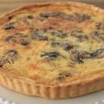 A baked quiche with a golden-brown crust is displayed on a scalloped white plate. The filling appears to include mushrooms and a creamy mixture, with visible browning on the surface, suggesting it is freshly baked and ready to serve.