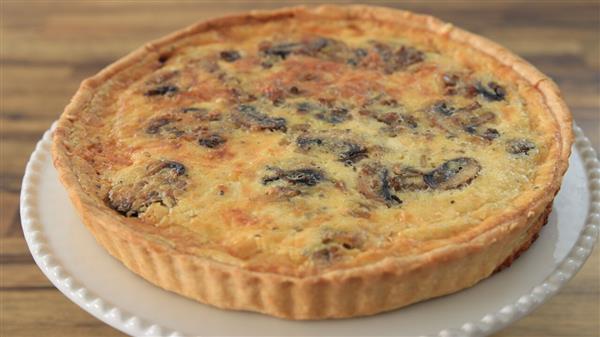 A baked quiche with a golden-brown crust is displayed on a scalloped white plate. The filling appears to include mushrooms and a creamy mixture, with visible browning on the surface, suggesting it is freshly baked and ready to serve.