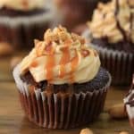 Chocolate cupcakes with creamy frosting topped with caramel drizzle and chopped nuts are displayed on a wooden surface. Some cupcakes also include a chocolate drizzle, and whole peanuts are scattered around the cupcakes.