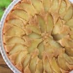 Top view of an apple upside-down cake on a white plate placed on a wooden surface. The cake is arranged with evenly distributed apple slices on top, glazed to a golden brown. Beside the plate are two green apples and one red apple.