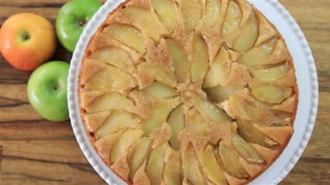 Top view of an apple upside-down cake on a white plate placed on a wooden surface. The cake is arranged with evenly distributed apple slices on top, glazed to a golden brown. Beside the plate are two green apples and one red apple.