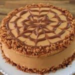 A round, caramel-colored cake sits on a white plate. The cake is decorated with a spiderweb pattern made of caramel drizzle on top. Crumbled cookie or nut pieces form a border around the edge of the cake's surface and at the bottom edge. The background is a wooden surface.