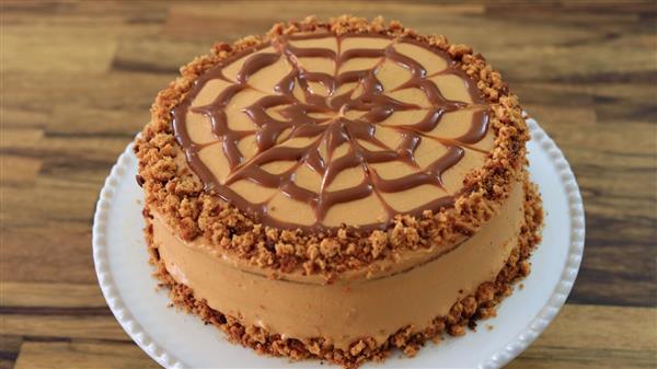 A round, caramel-colored cake sits on a white plate. The cake is decorated with a spiderweb pattern made of caramel drizzle on top. Crumbled cookie or nut pieces form a border around the edge of the cake's surface and at the bottom edge. The background is a wooden surface.