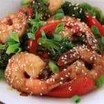A white bowl filled with a colorful shrimp stir-fry. The dish includes plump shrimp, broccoli florets, and bright red bell pepper slices. The stir-fry is garnished with sesame seeds and chopped green onions. All ingredients are coated in a savory sauce.
