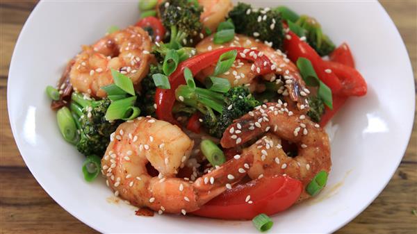 A white bowl filled with a colorful shrimp stir-fry. The dish includes plump shrimp, broccoli florets, and bright red bell pepper slices. The stir-fry is garnished with sesame seeds and chopped green onions. All ingredients are coated in a savory sauce.