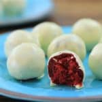 A close-up of a blue plate with multiple white chocolate-covered red velvet cake balls. One of the cake balls is cut in half, revealing the vibrant red interior. Another plate with more cake balls is slightly blurred in the background.