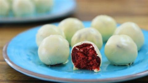 A close-up of a blue plate with multiple white chocolate-covered red velvet cake balls. One of the cake balls is cut in half, revealing the vibrant red interior. Another plate with more cake balls is slightly blurred in the background.