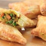 Golden, flaky vegetable puff pastries are arranged on a wooden surface. One pastry is split open, revealing a filling of mixed vegetables, including peas and carrots, in a savory sauce.