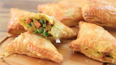 Golden, flaky vegetable puff pastries are arranged on a wooden surface. One pastry is split open, revealing a filling of mixed vegetables, including peas and carrots, in a savory sauce.